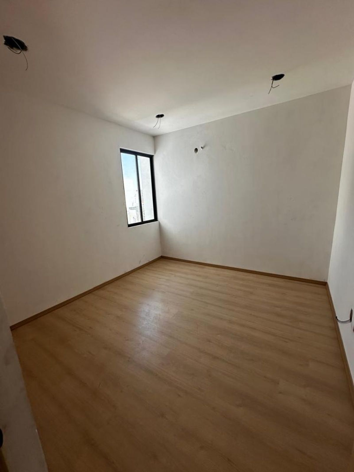 Habitación vacía con piso de madera clara, paredes blancas y ventana con marco negro