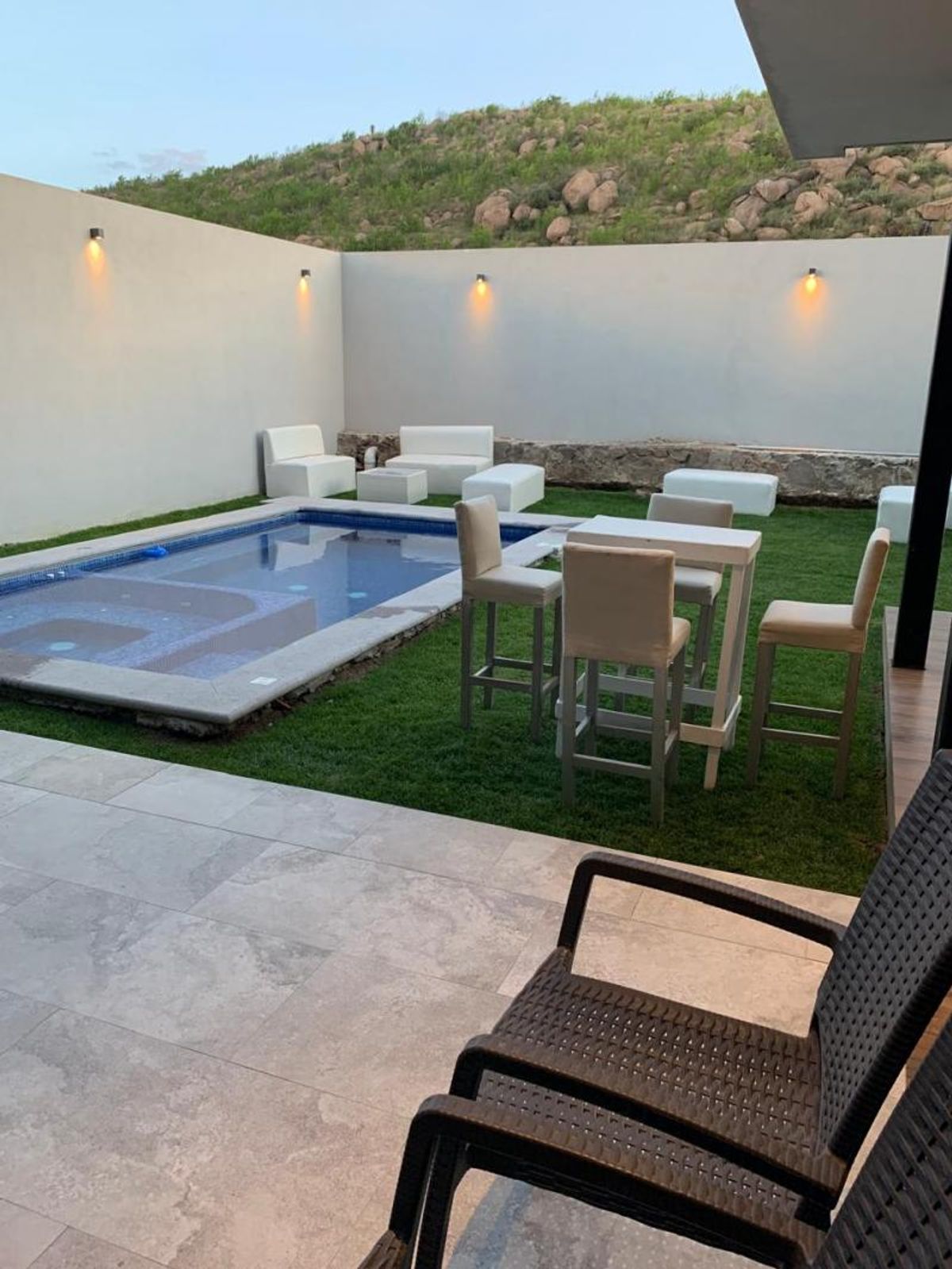 Jardín posterior con alberca, césped, mesa alta con bancos, sillones blancos y terraza