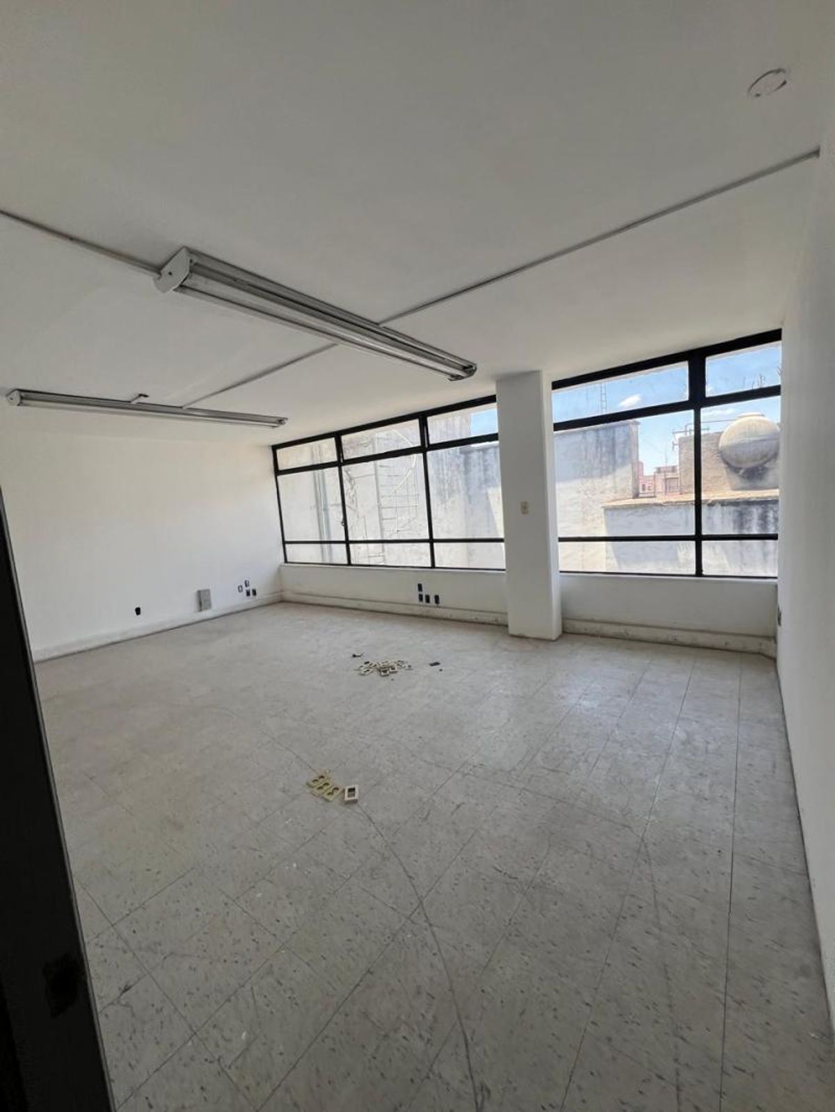 Espacio vacío con paredes blancas, piso claro, luminarias alargadas y ventanales con marco negro