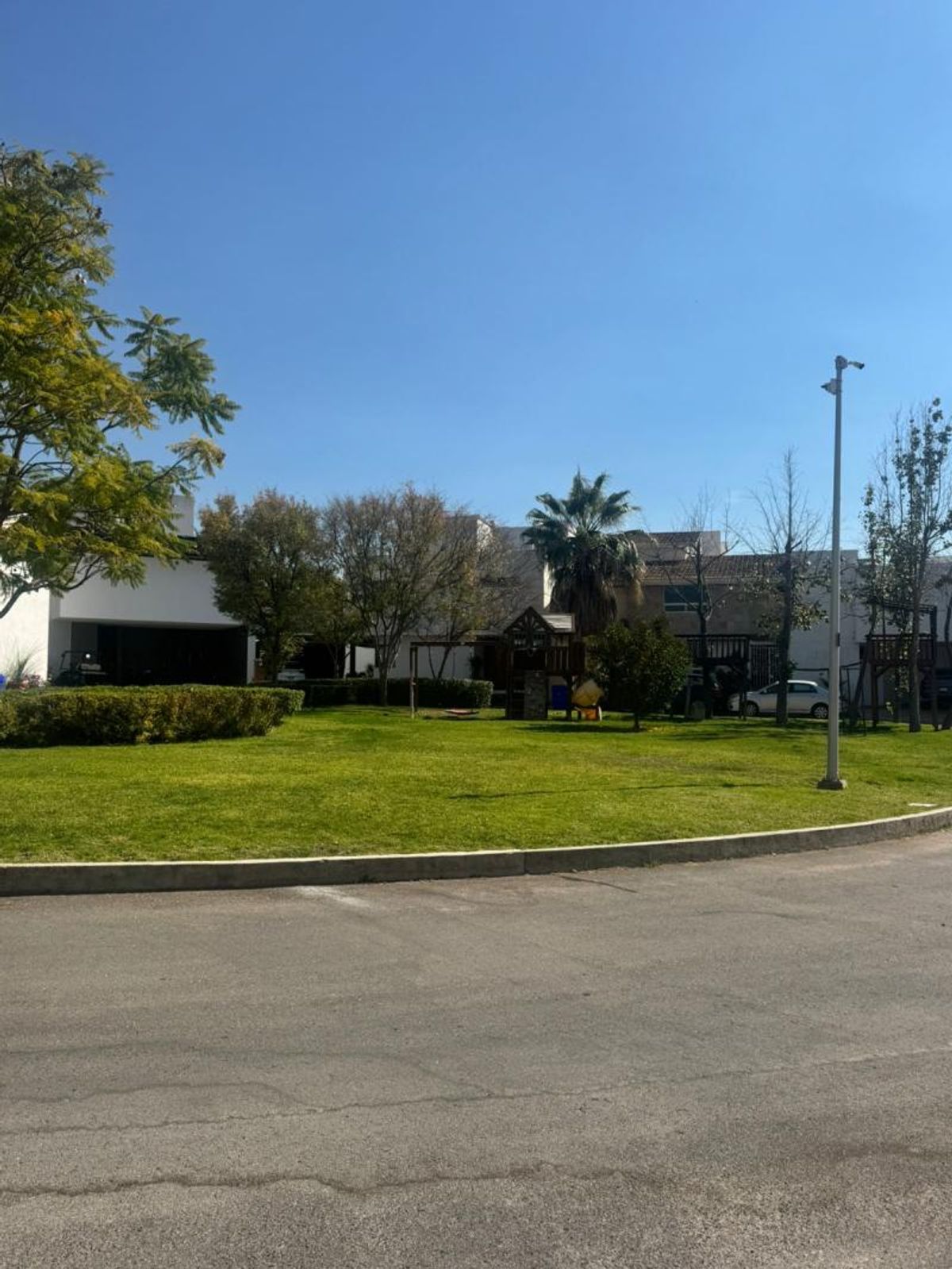 Jardín comunitario con pasto verde, árboles, juegos infantiles de madera y casas al fondo