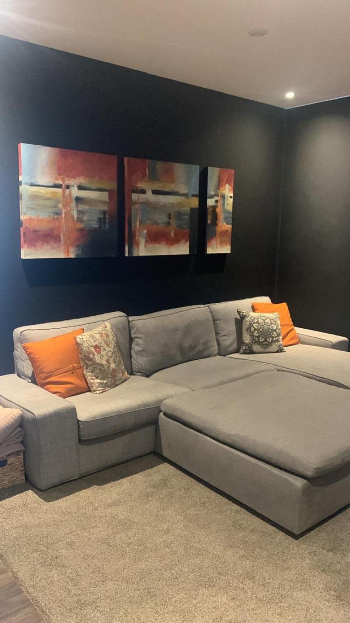 Sala con paredes oscuras, sofá seccional gris con cojines naranjas, alfombra y cuadro decorativo