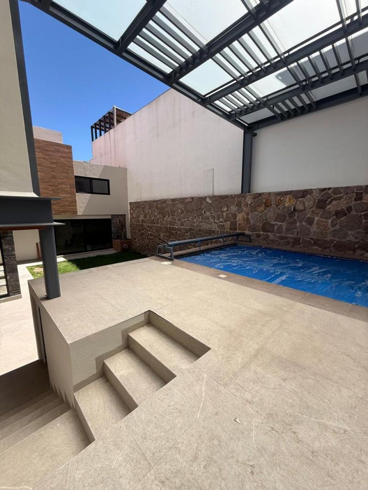 Terraza exterior con alberca, pared de piedra, piso claro y pergolado de cristal