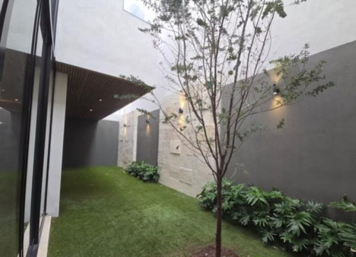 Jardín lateral con césped sintético, árbol joven, plantas de follaje y muro iluminado
