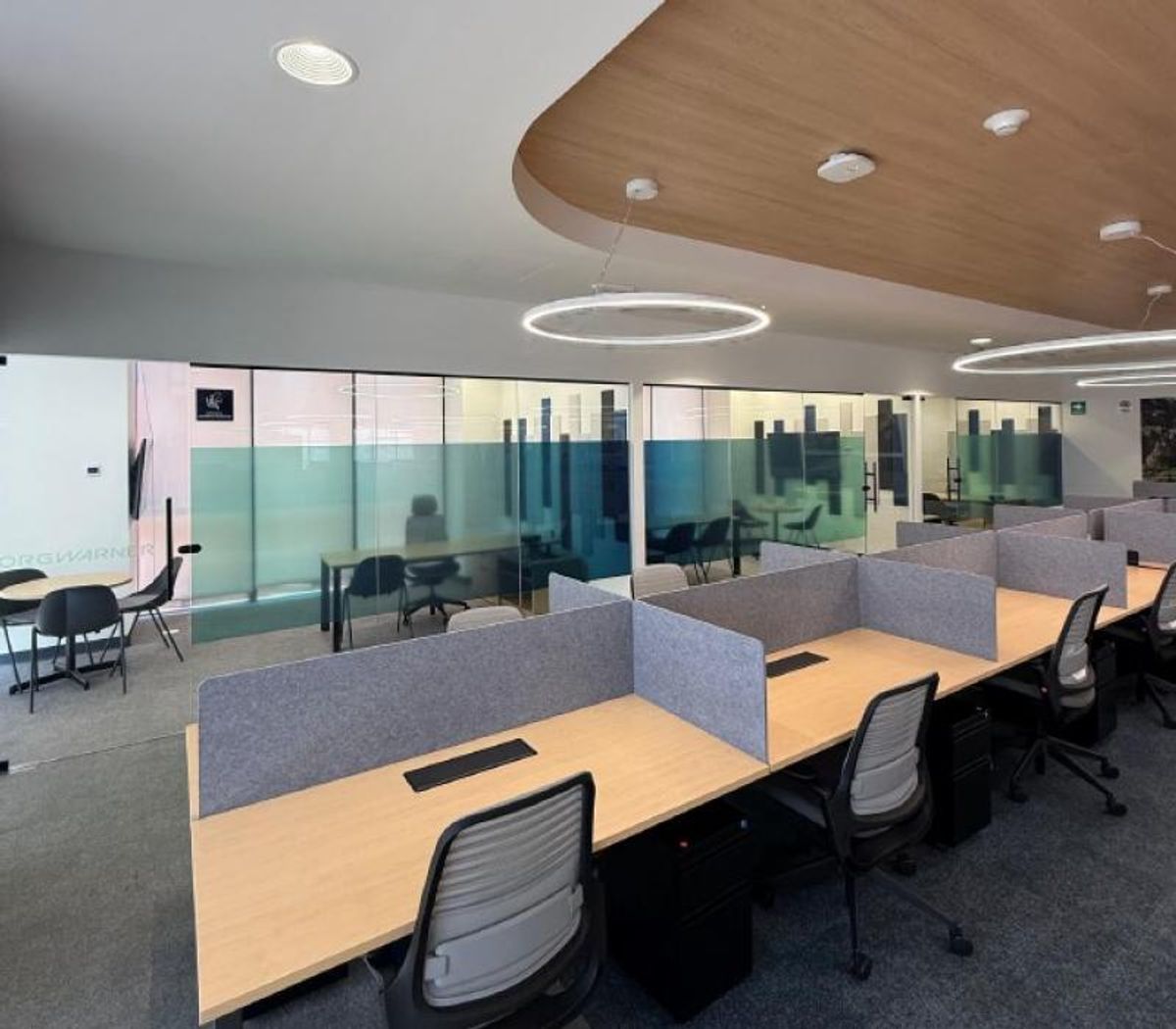 Área de trabajo con escritorios de madera, divisores grises, sillas ergonómicas y mamparas de cristal