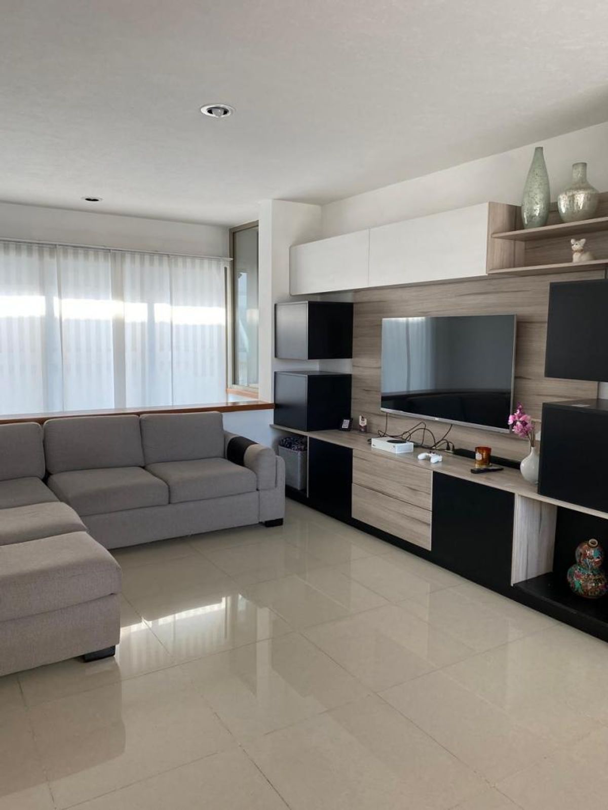 Sala de estar con sofá gris esquinero, piso cerámico brillante y mueble de televisión de madera