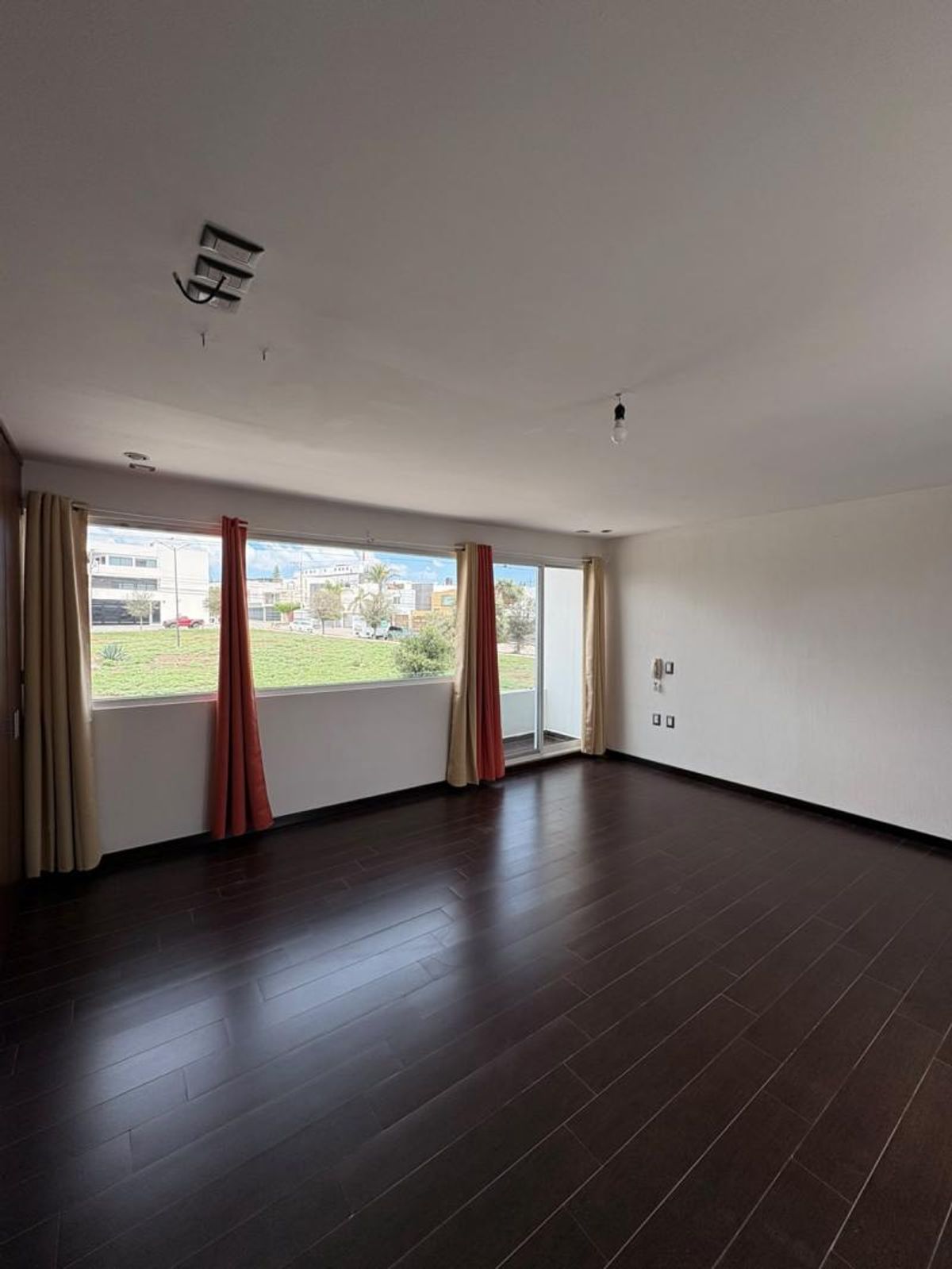 Habitación vacía con piso de madera oscura, paredes blancas y ventanales hacia área verde