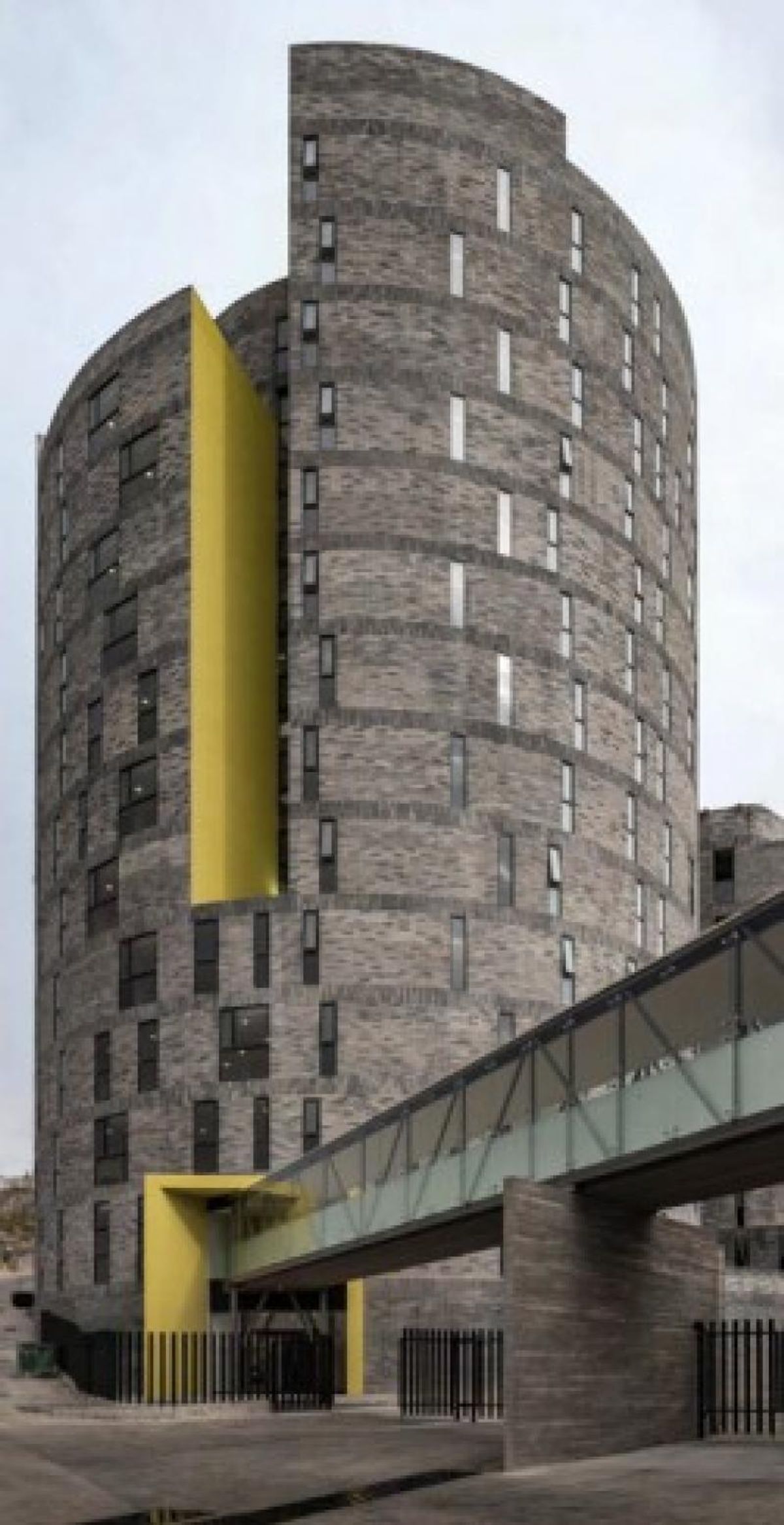 Torre curva de ladrillo gris con detalles amarillos y puente peatonal elevado