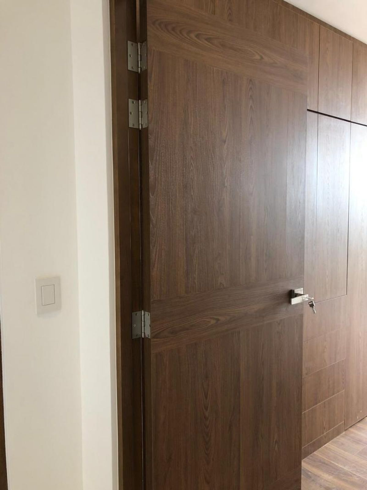 Puerta de madera oscura con bisagras metálicas, manija plateada y pared blanca con apagador