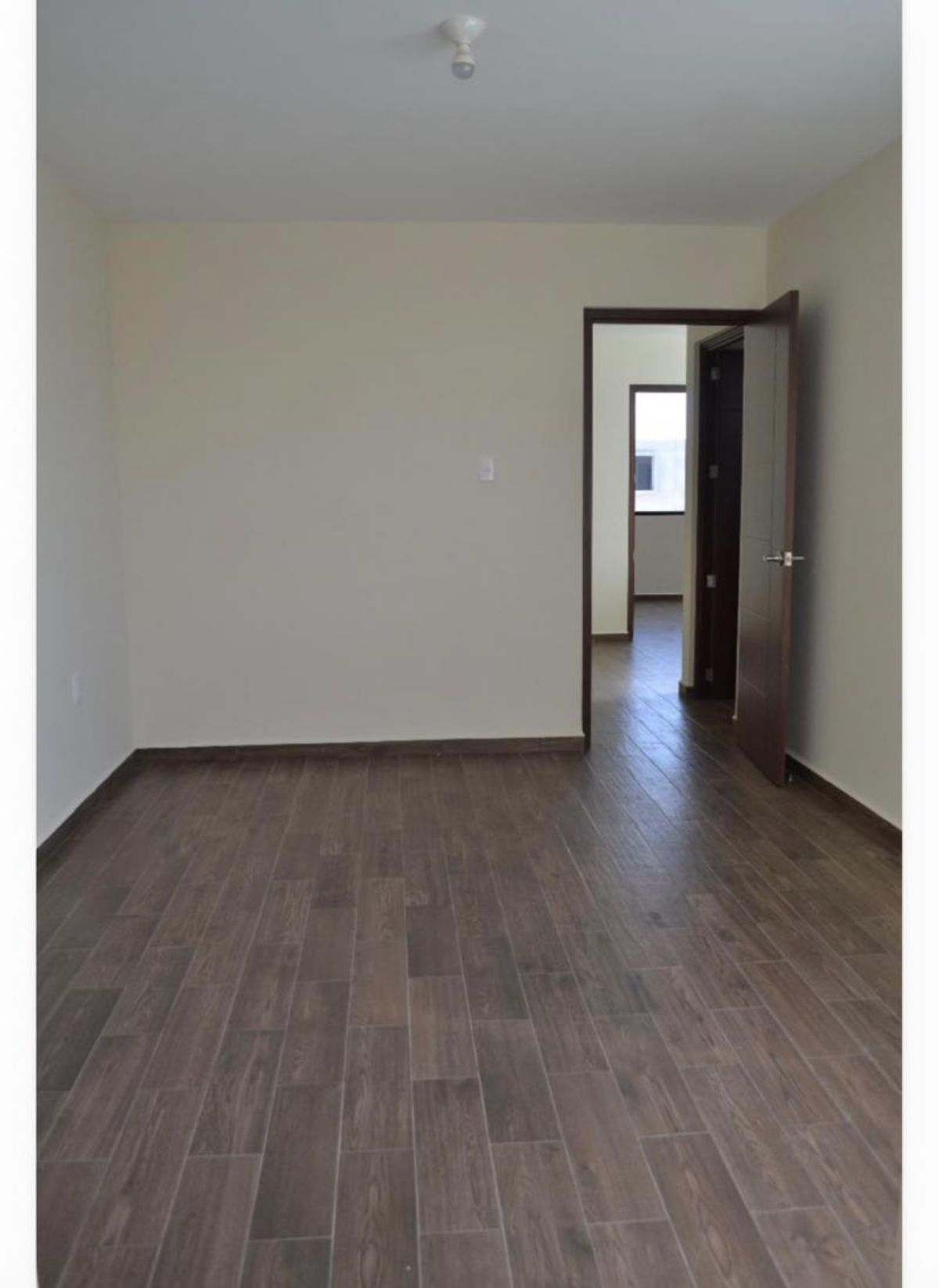 Habitación vacía con piso cerámico tipo madera, paredes claras y puerta de madera abierta