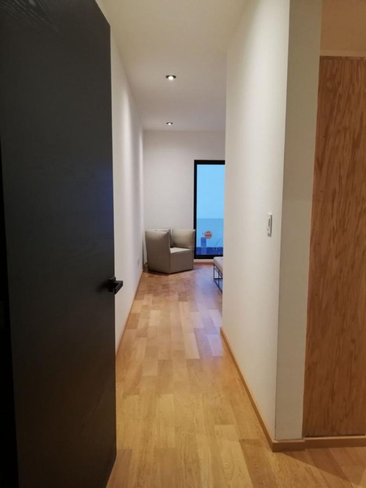 Pasillo con piso de madera clara, puerta oscura, paredes blancas y sillón gris
