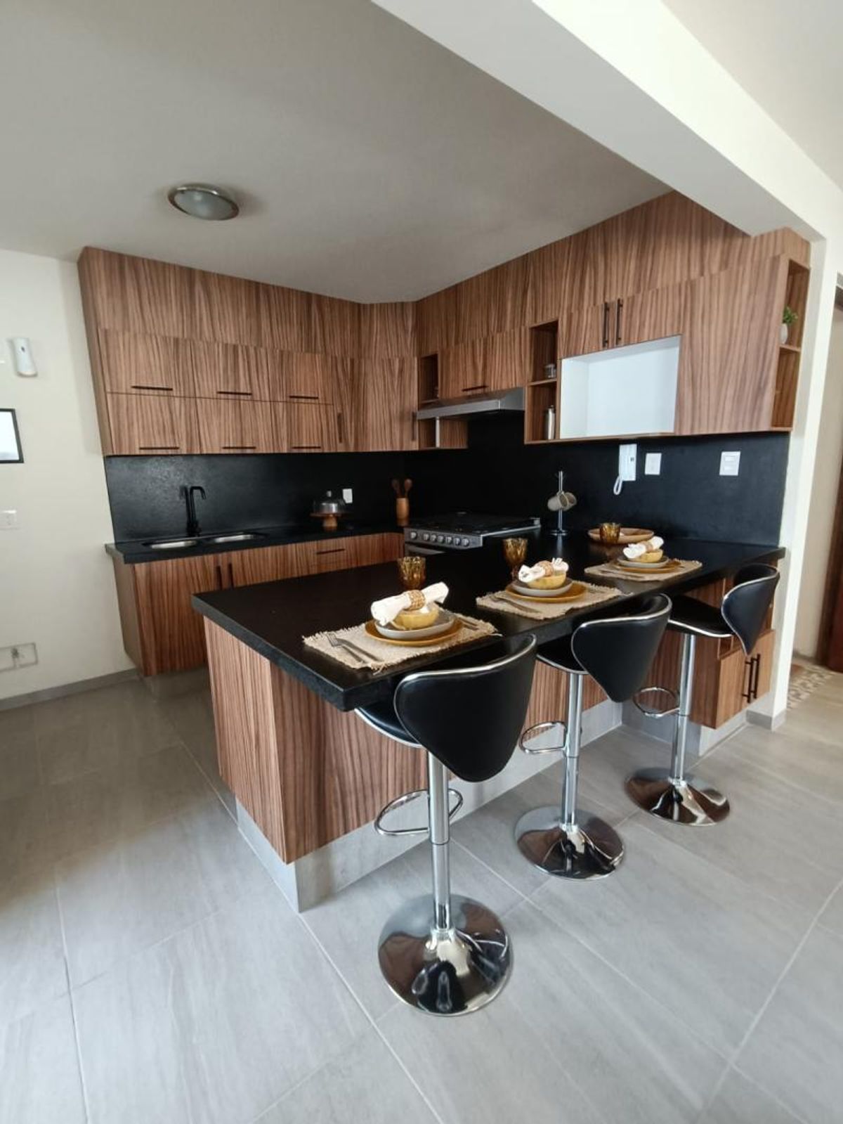 Cocina integral con gabinetes de madera, cubiertas negras y barra desayunadora con tres bancos