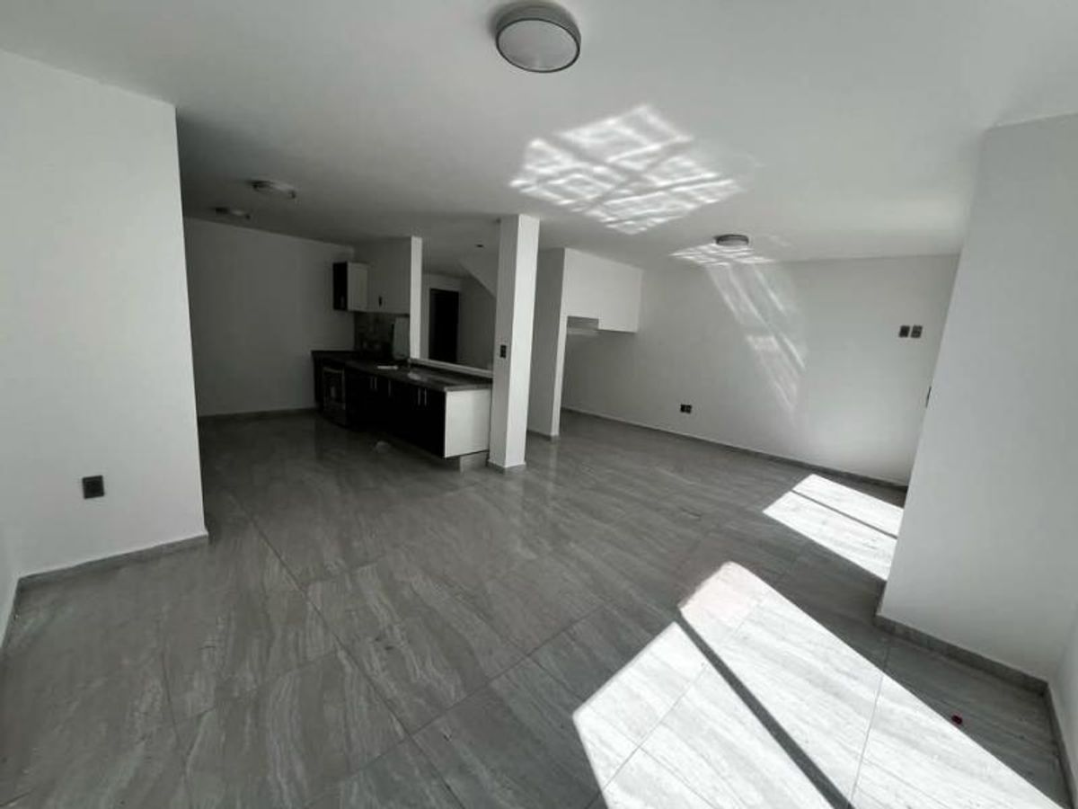 Espacio abierto con piso cerámico gris, luz natural, paredes blancas y cocina al fondo