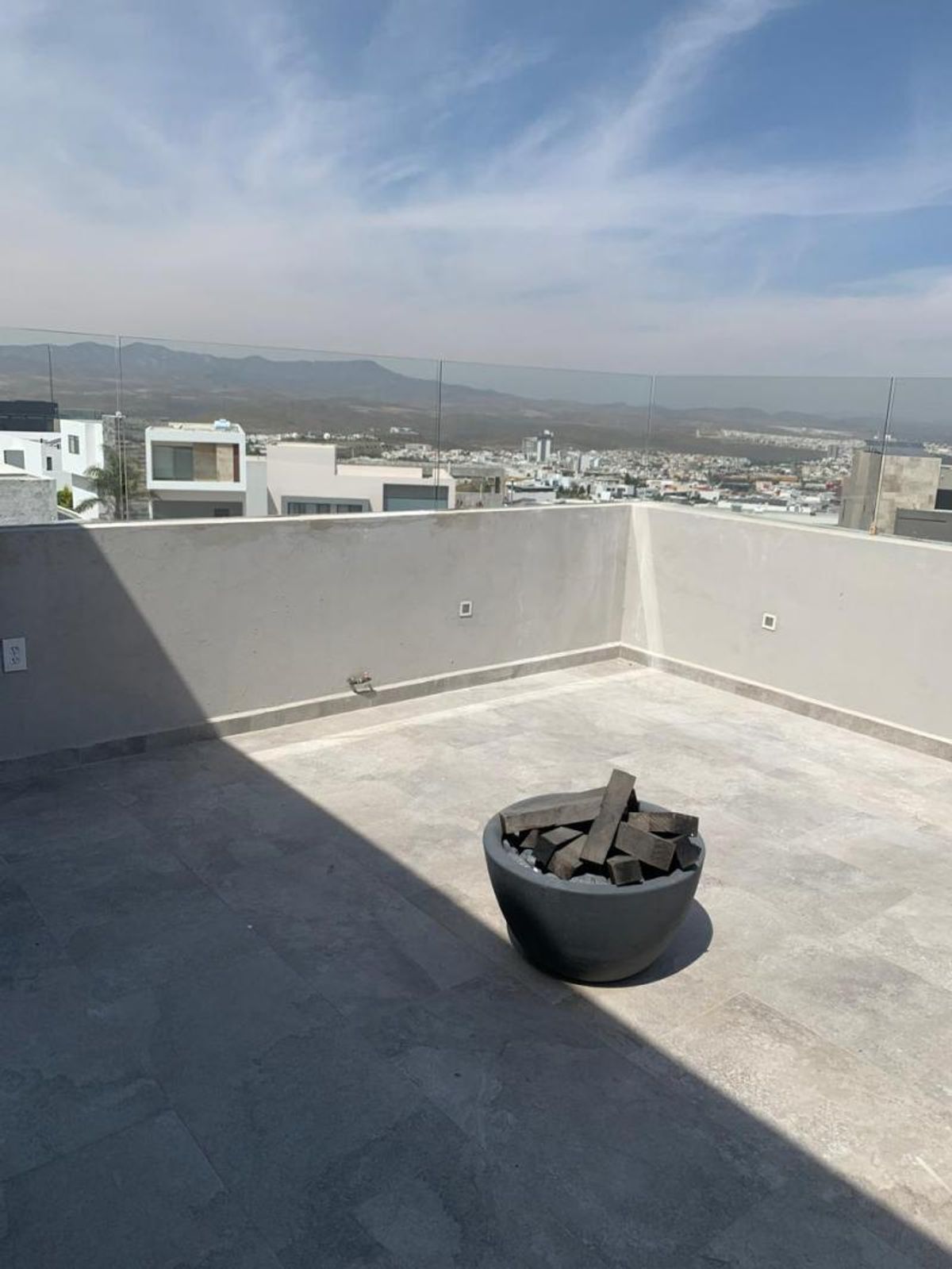 Terraza con piso gris, fogatero circular, barandal de cristal y vista a la ciudad