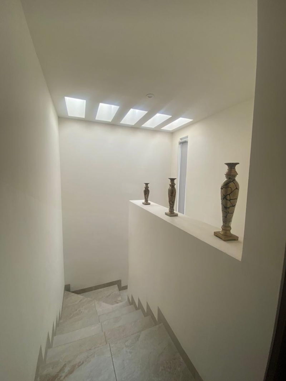 Escalera interior de tonos claros con tragaluces y jarrones sobre un medio muro