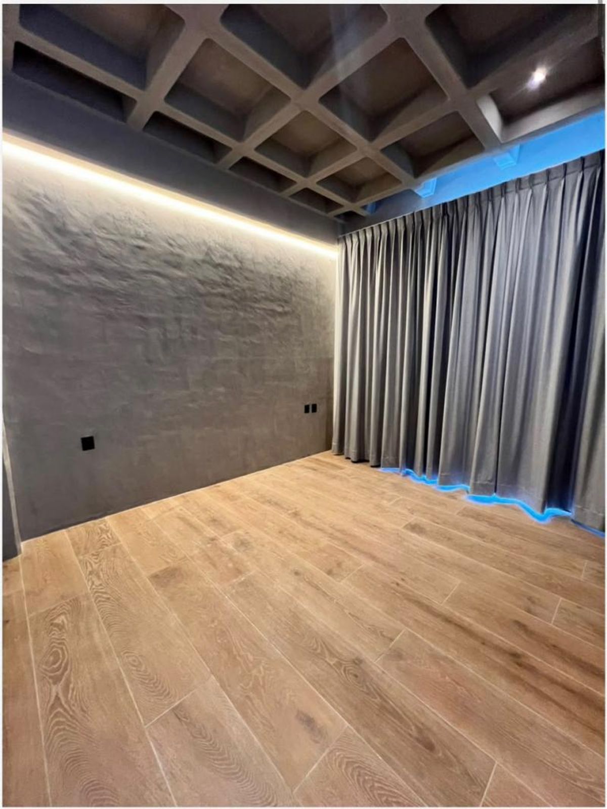 Habitación vacía con piso de madera, pared gris texturizada, cortinas y techo reticulado