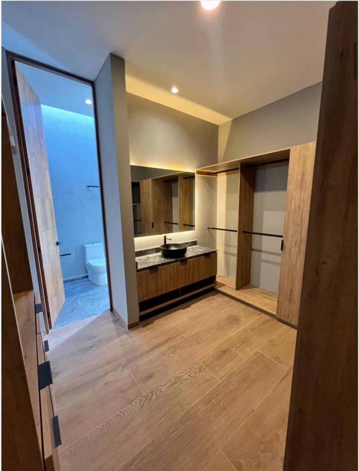 Vestidor de madera con lavabo negro sobre cubierta, espejo iluminado y acceso al baño