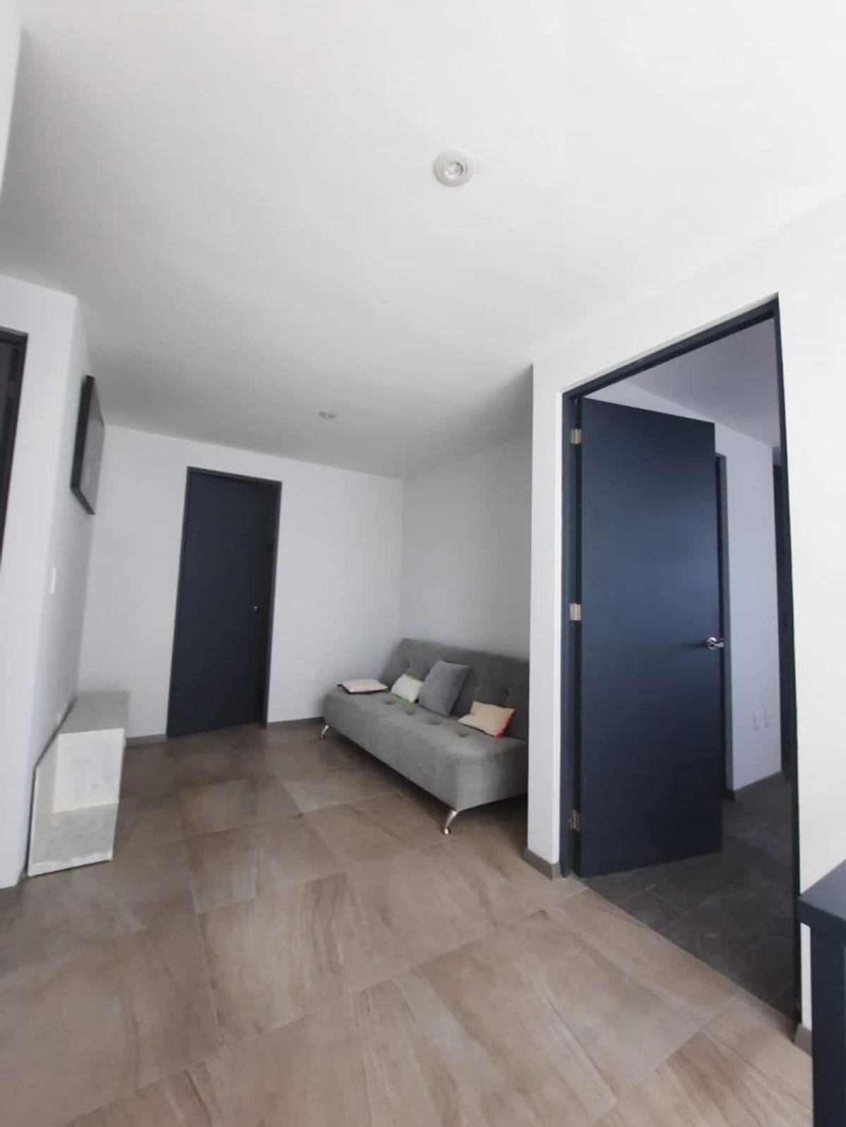 Sala con sofá gris, cojines decorativos, paredes blancas, piso cerámico y puertas oscuras
