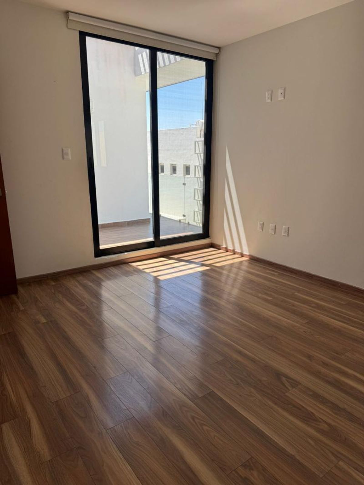 Habitación vacía con piso laminado de madera, paredes claras y puerta corrediza de cristal