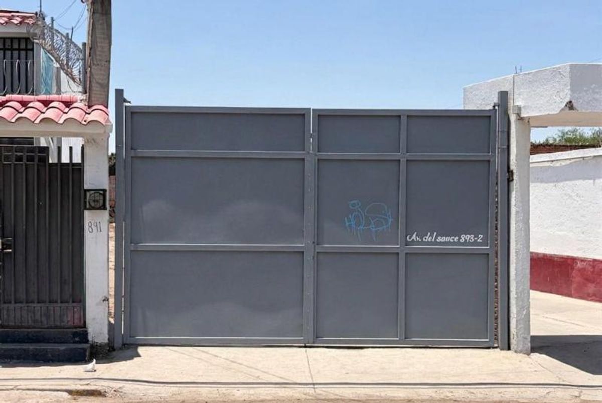 Portón metálico gris con dirección pintada entre muros blancos sobre banqueta de concreto