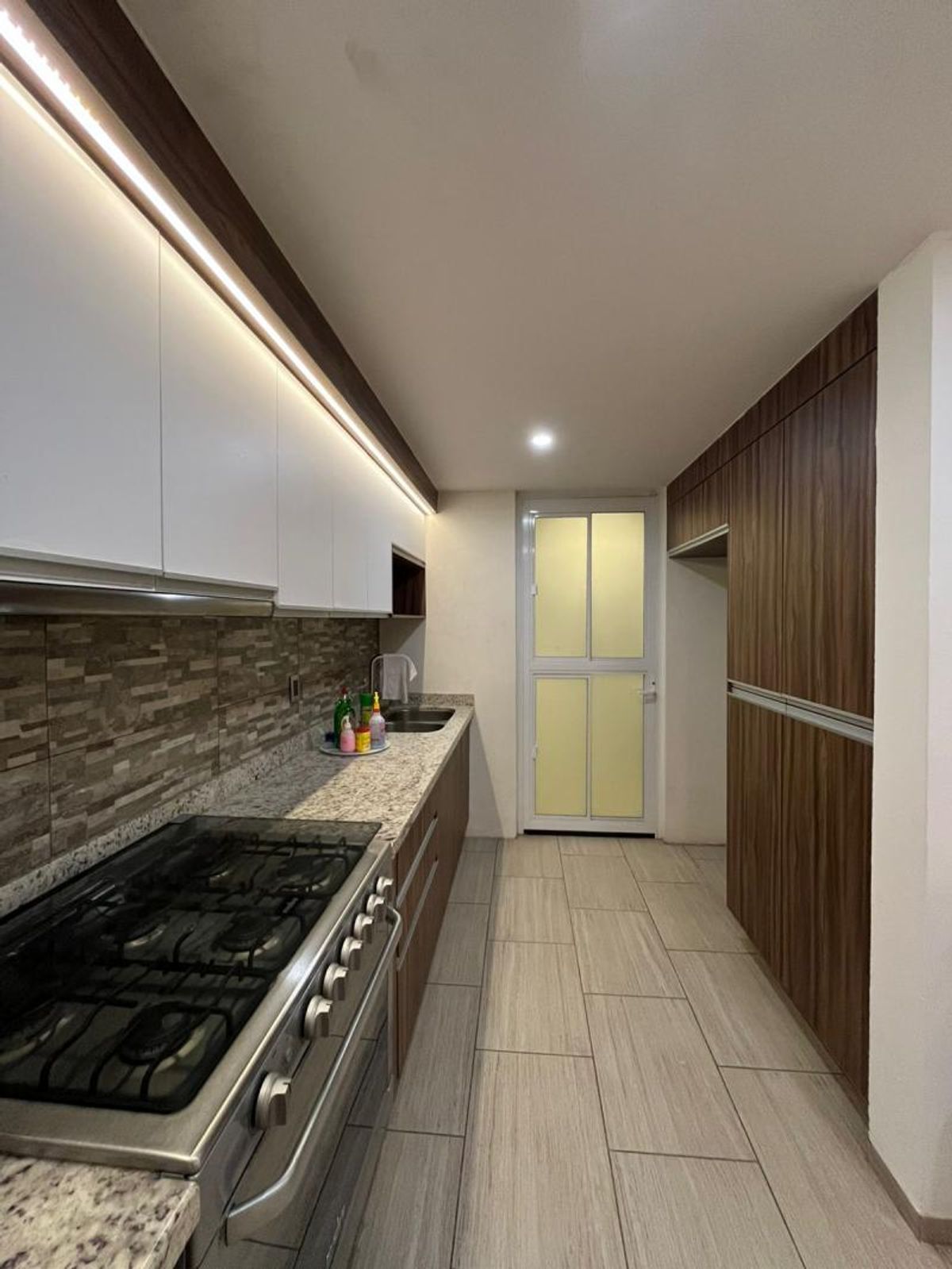 Cocina alargada con gabinetes blancos, iluminación LED, cubierta de granito y alacena de madera
