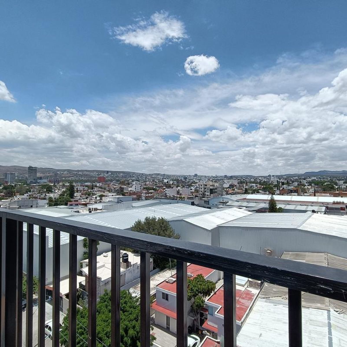 Vista de la ciudad desde un balcón con barandal de metal negro bajo cielo azul nublado