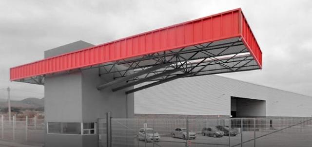 Caseta de acceso con gran marquesina roja metálica y nave industrial gris al fondo