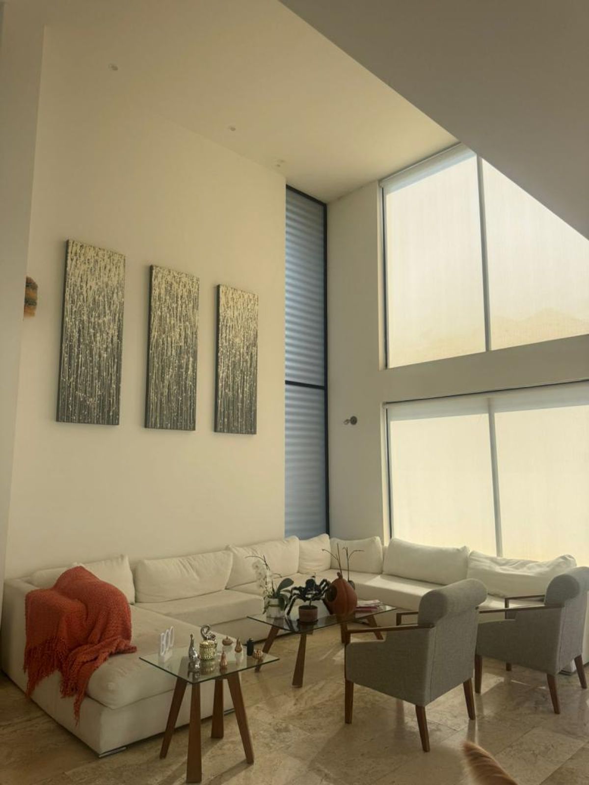 Sala de doble altura con sofá modular claro, sillones grises, ventanales y cuadros decorativos