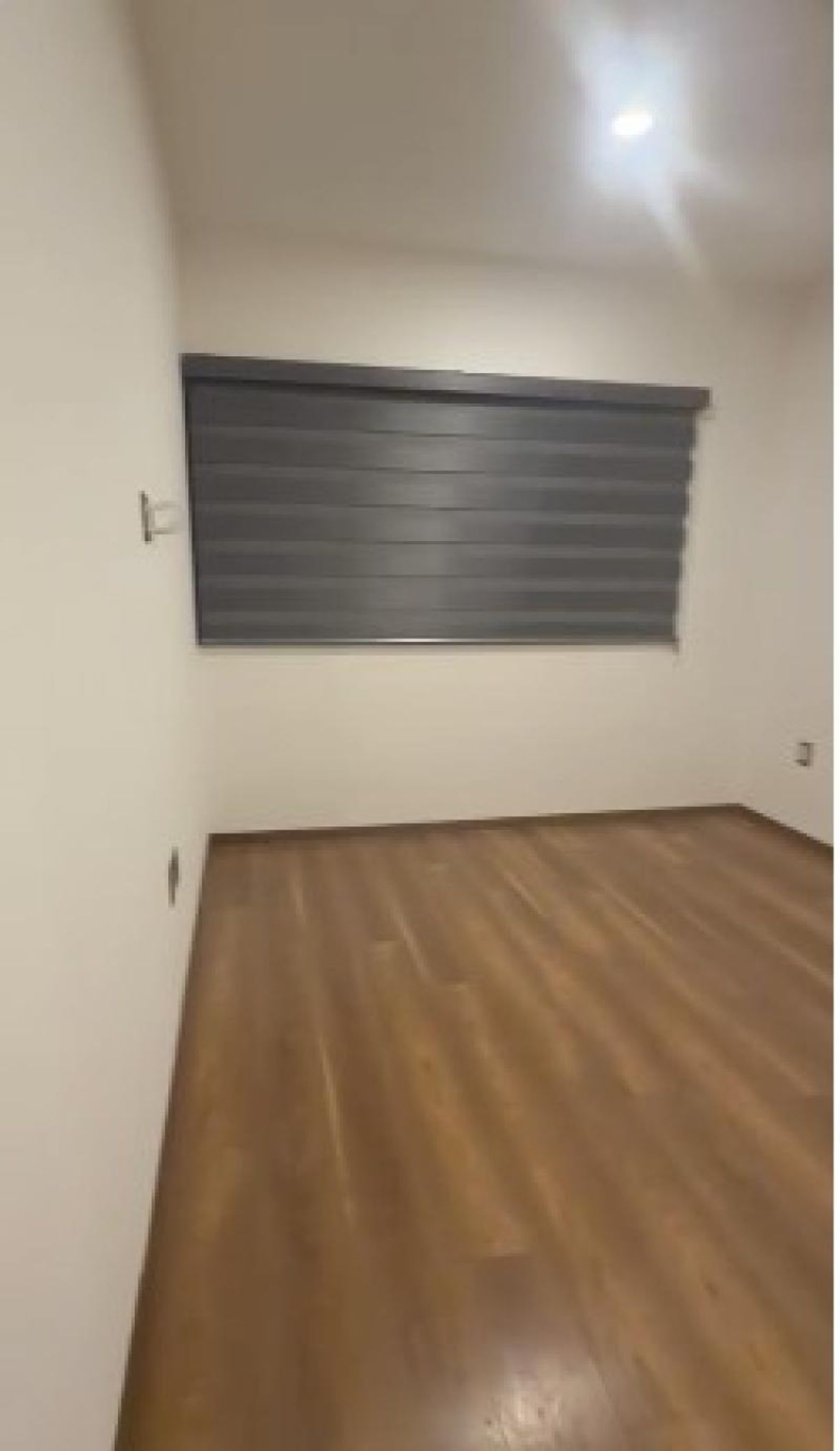 Habitación vacía con piso de madera, paredes blancas y ventana con persiana gris