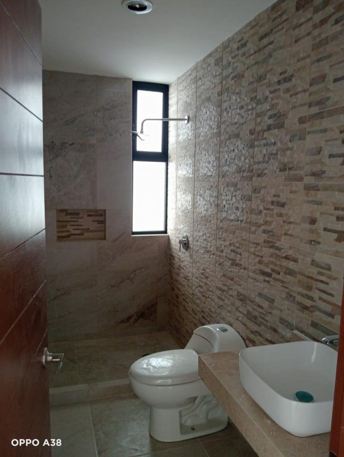 Baño con inodoro blanco, lavabo sobre cubierta de piedra y regadera al fondo con ventana