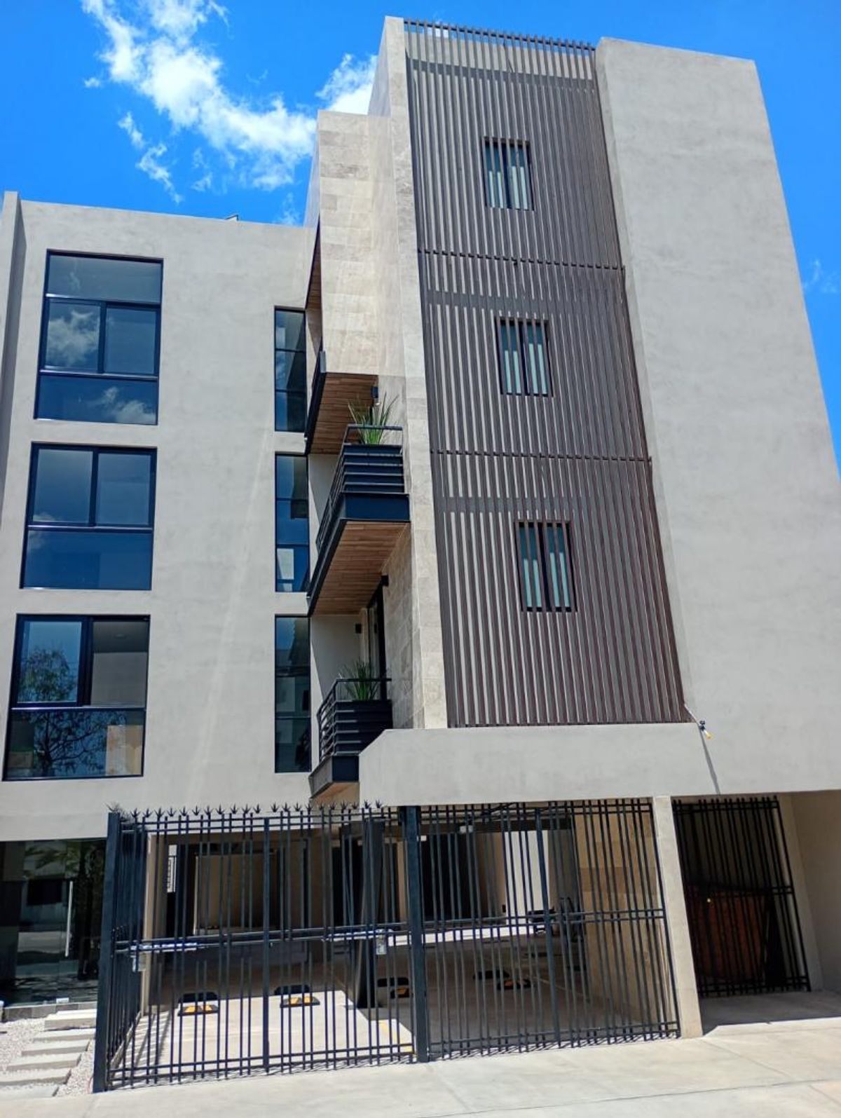 Edificio moderno de departamentos con fachada gris, paneles verticales, balcones y portón metálico negro