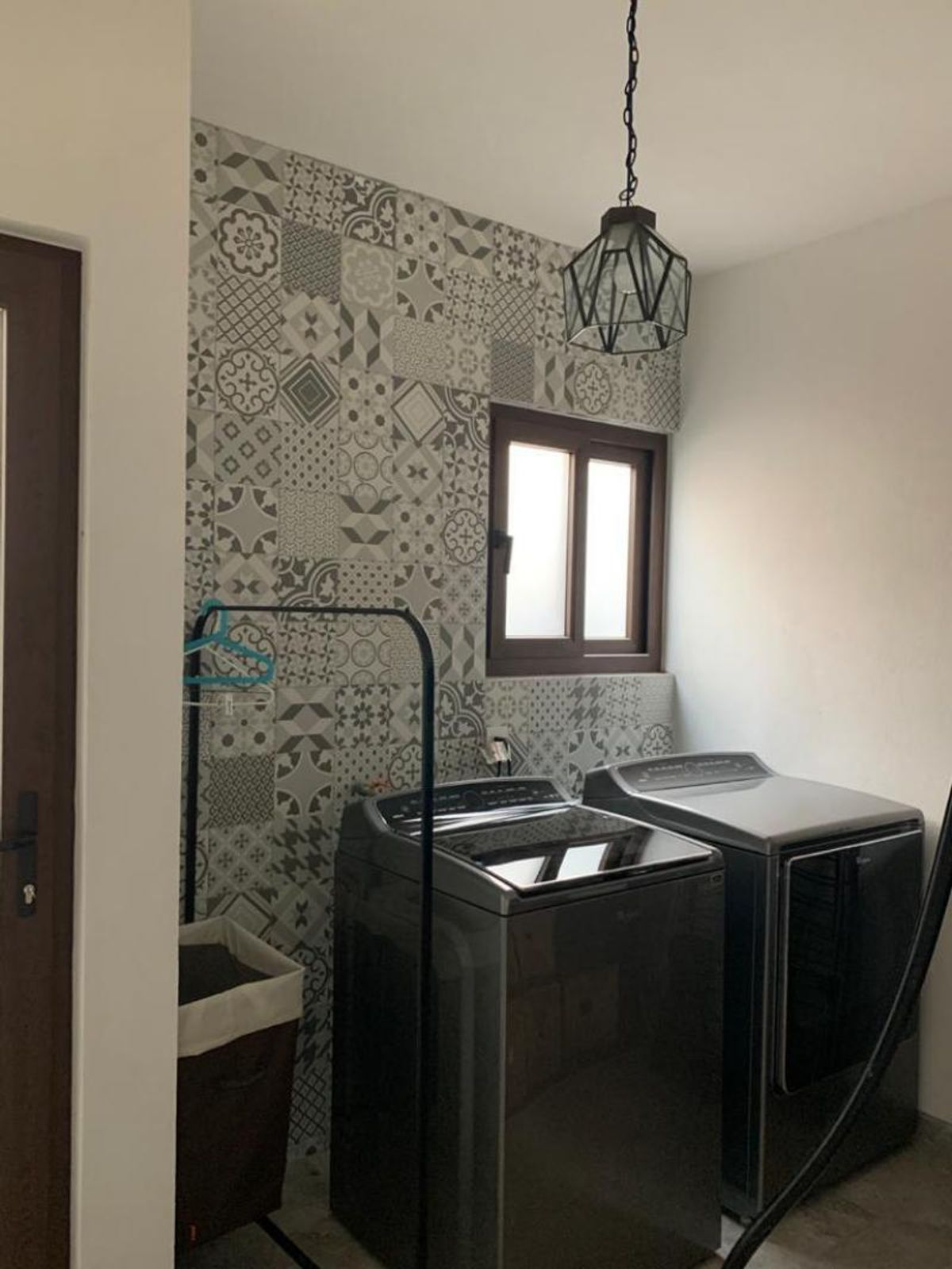 Área de lavado con lavadora, secadora, pared de azulejos estampados y lámpara colgante negra