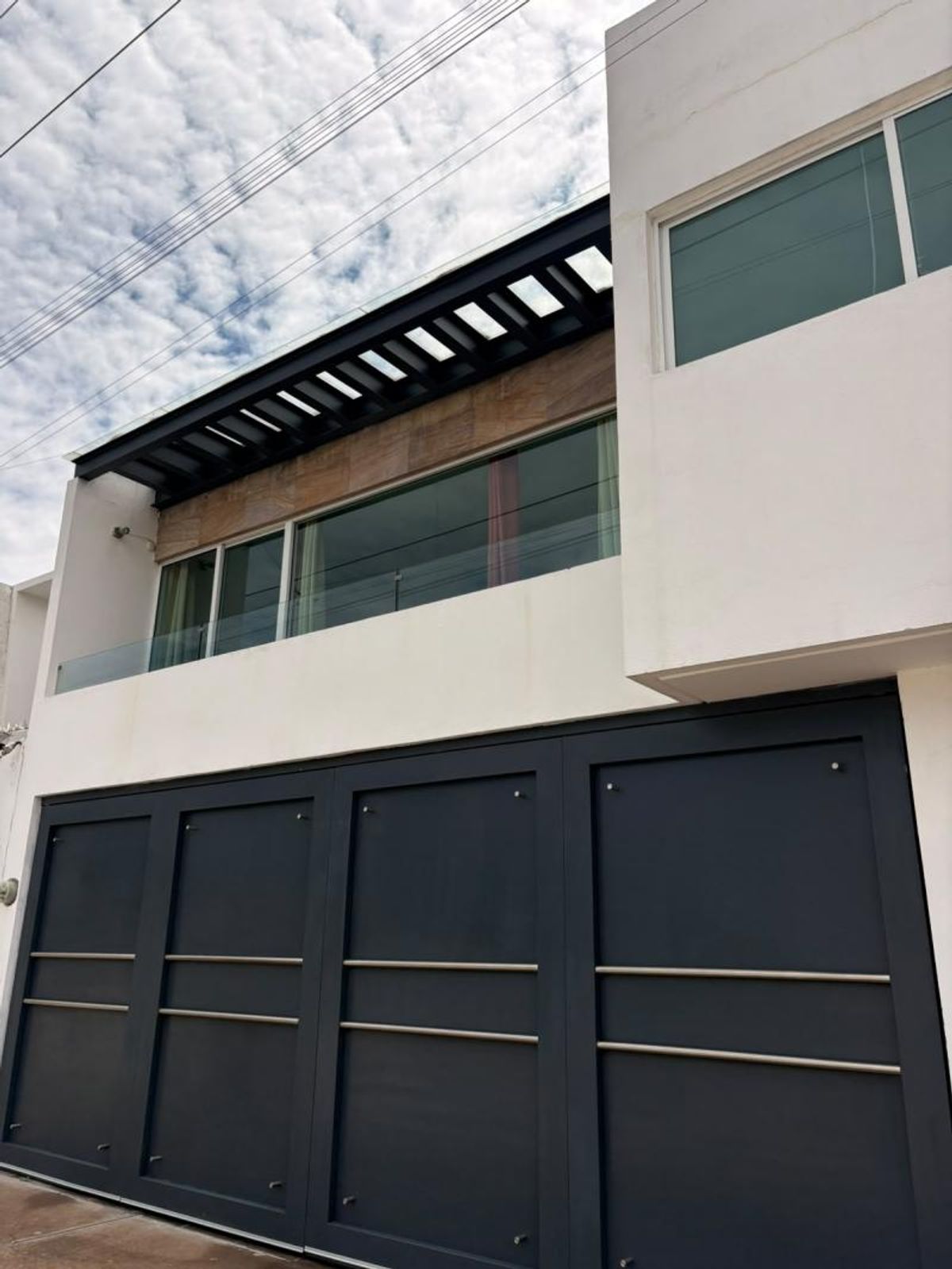 Fachada moderna con portón gris oscuro, muros blancos y ventanales con pérgola metálica