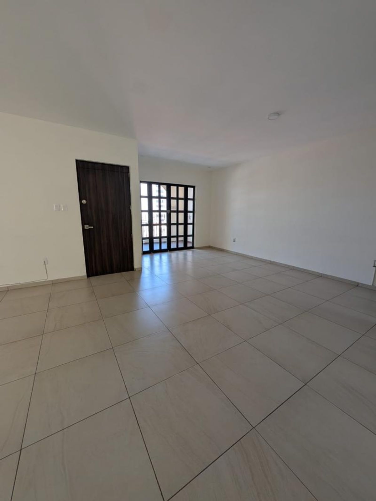 Sala vacía con piso de loseta clara, puerta de madera oscura y ventanal al fondo