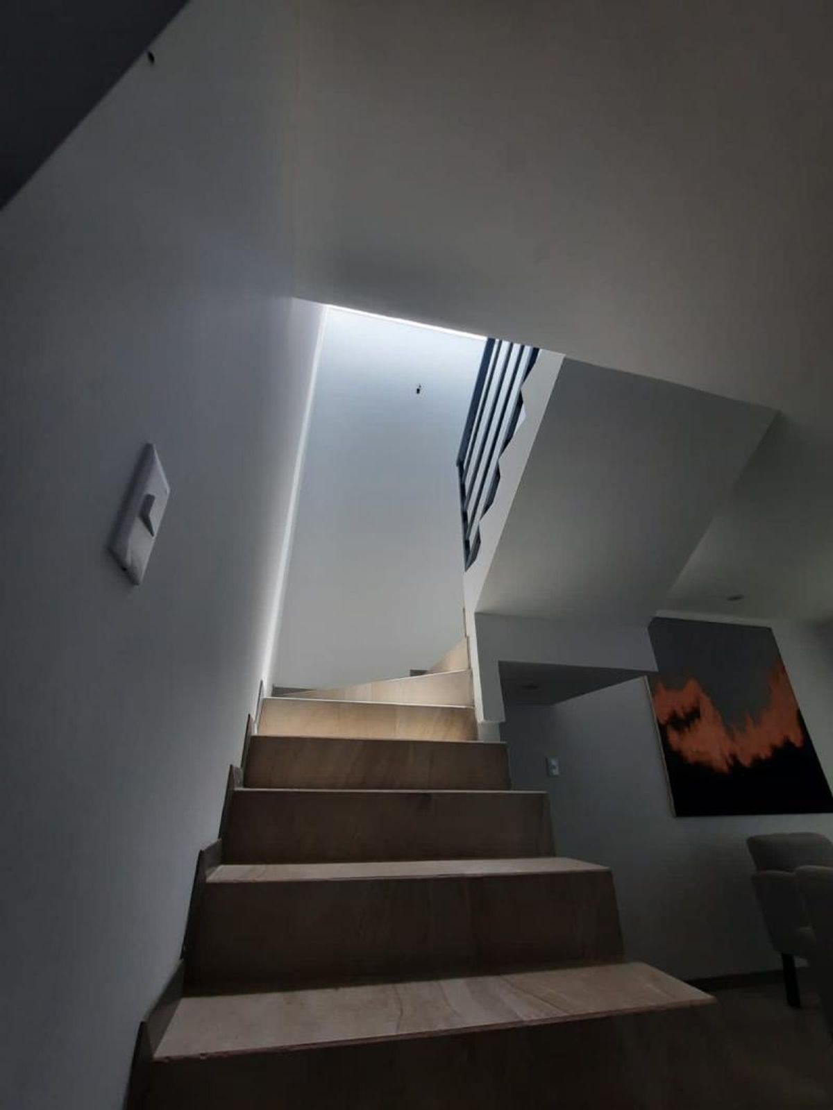 Escalera de madera iluminada por tragaluz superior entre paredes blancas y barandal metálico negro