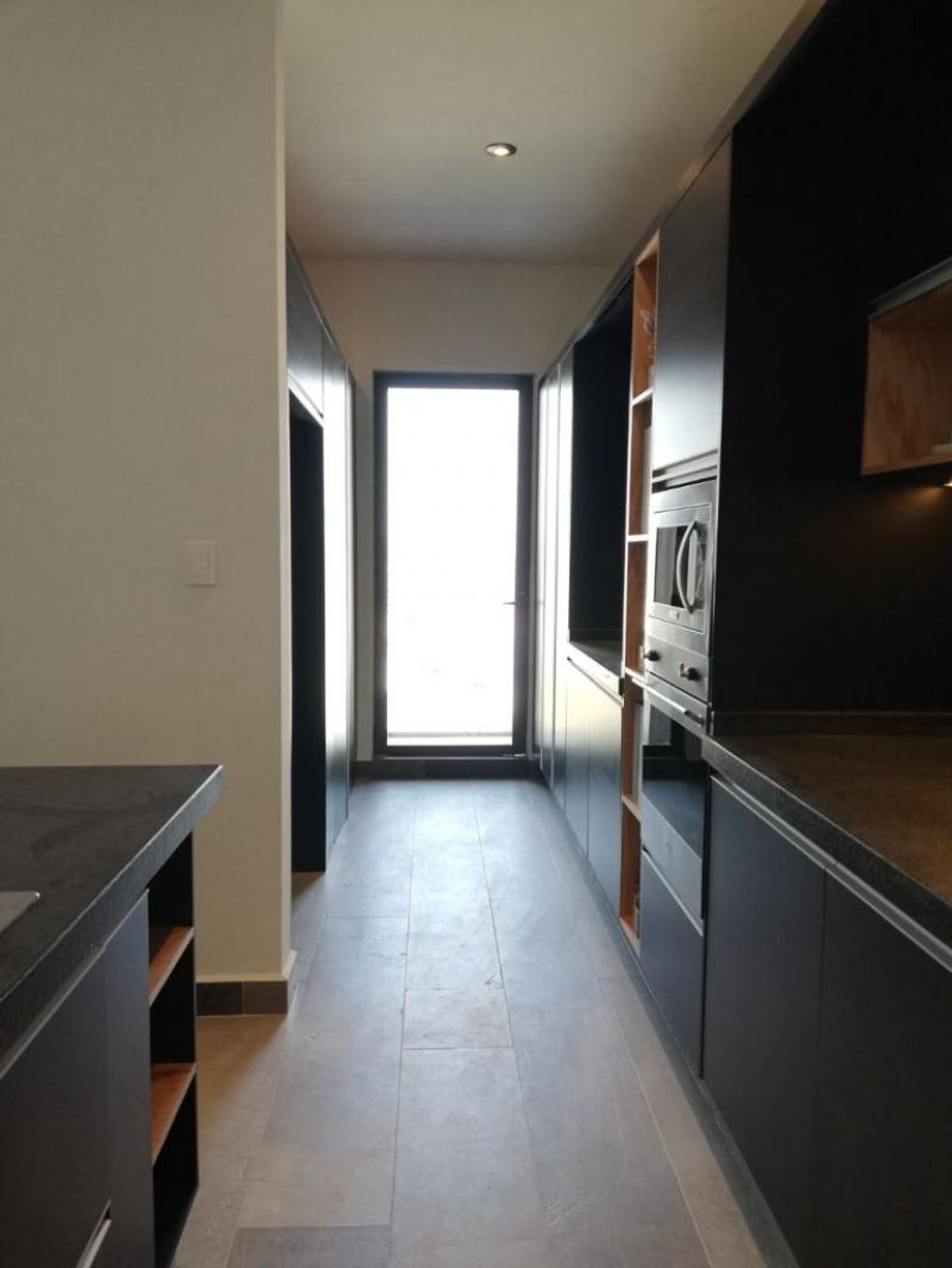 Cocina tipo pasillo con gabinetes oscuros, hornos empotrados, piso claro y ventana al fondo