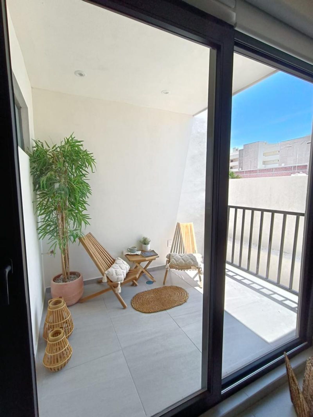 Balcón con piso gris, sillas de madera, mesa auxiliar, planta en maceta y ventanal