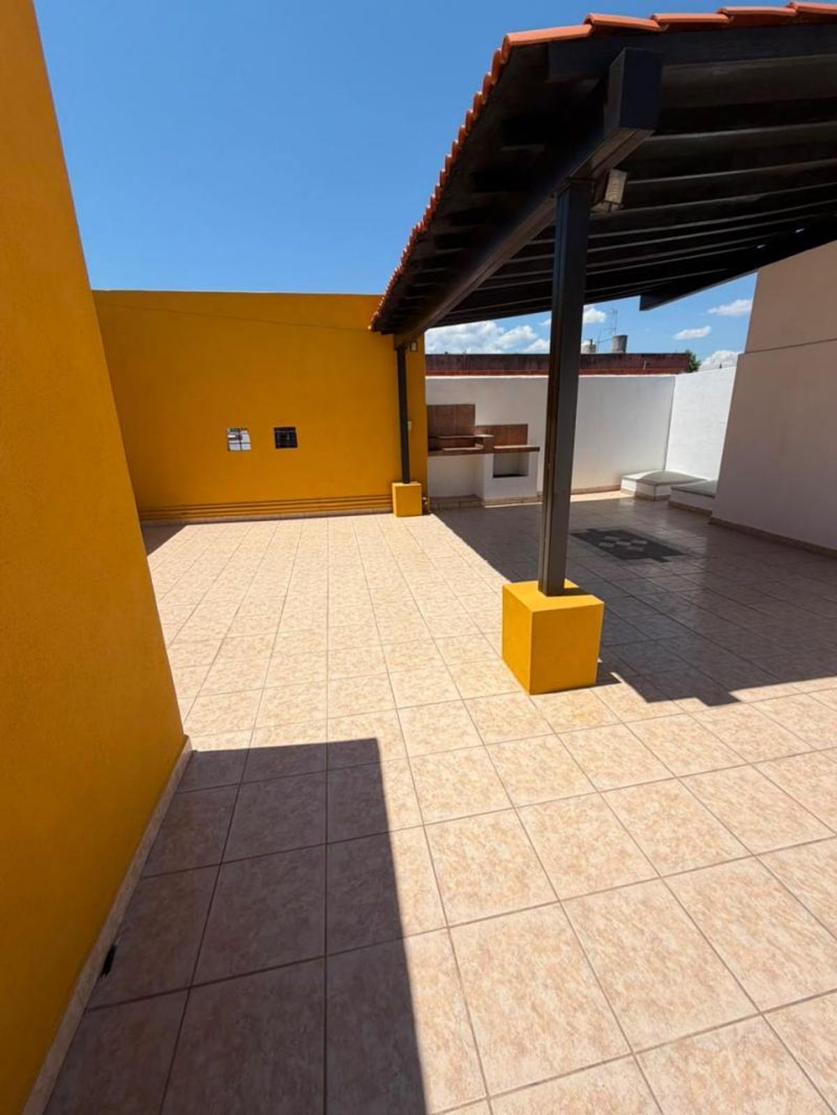 Patio exterior con piso de azulejo beige, muros amarillos y estructura techada con asador