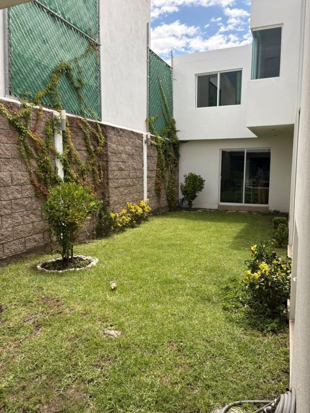 Jardín con pasto verde, arbustos y muro de bloques junto a la casa blanca