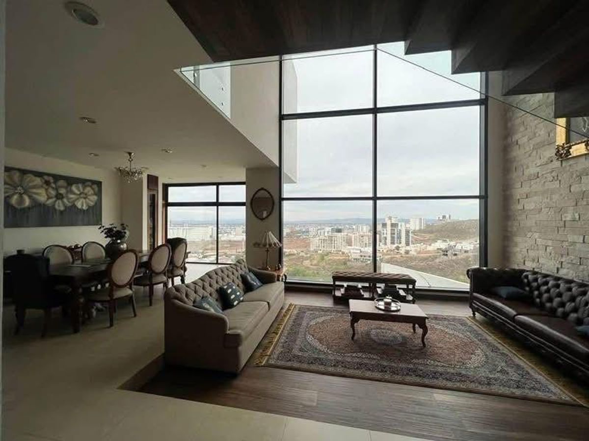 Sala y comedor de doble altura con ventanales, sofás y vista a la ciudad