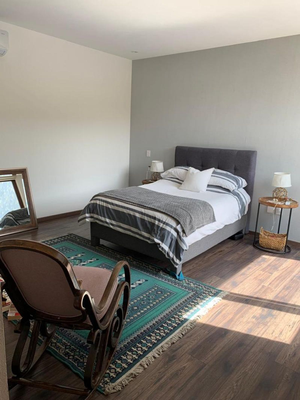 Recámara con cama tapizada gris, mecedora de madera y tapete verde en piso laminado