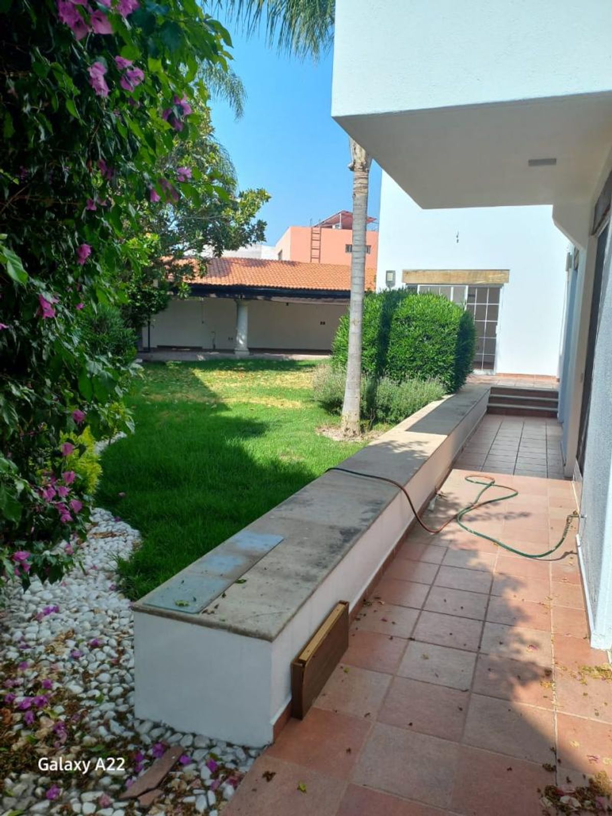 Jardín con césped verde, pasillo con baldosas de terracota y terraza cubierta al fondo