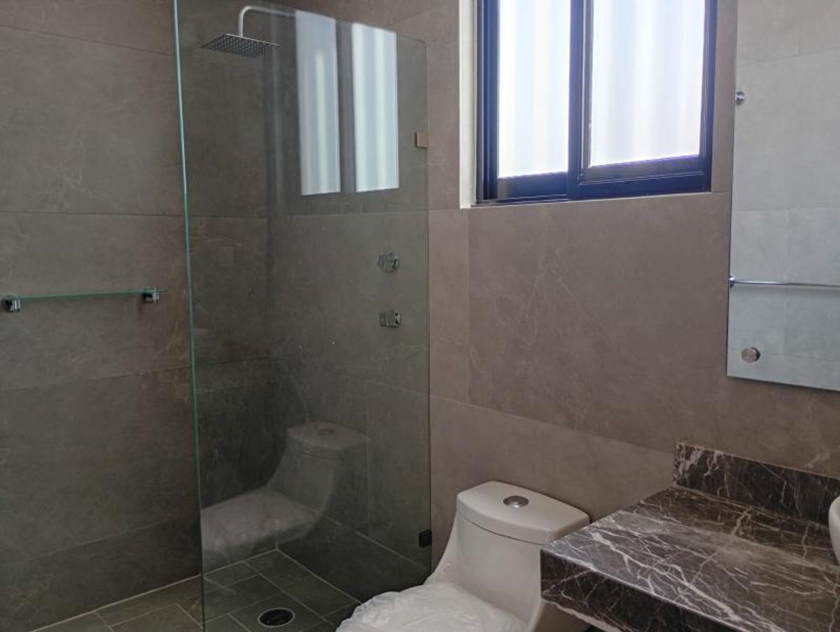 Baño con cancel de cristal, inodoro blanco, muros revestidos en gris y cubierta de mármol