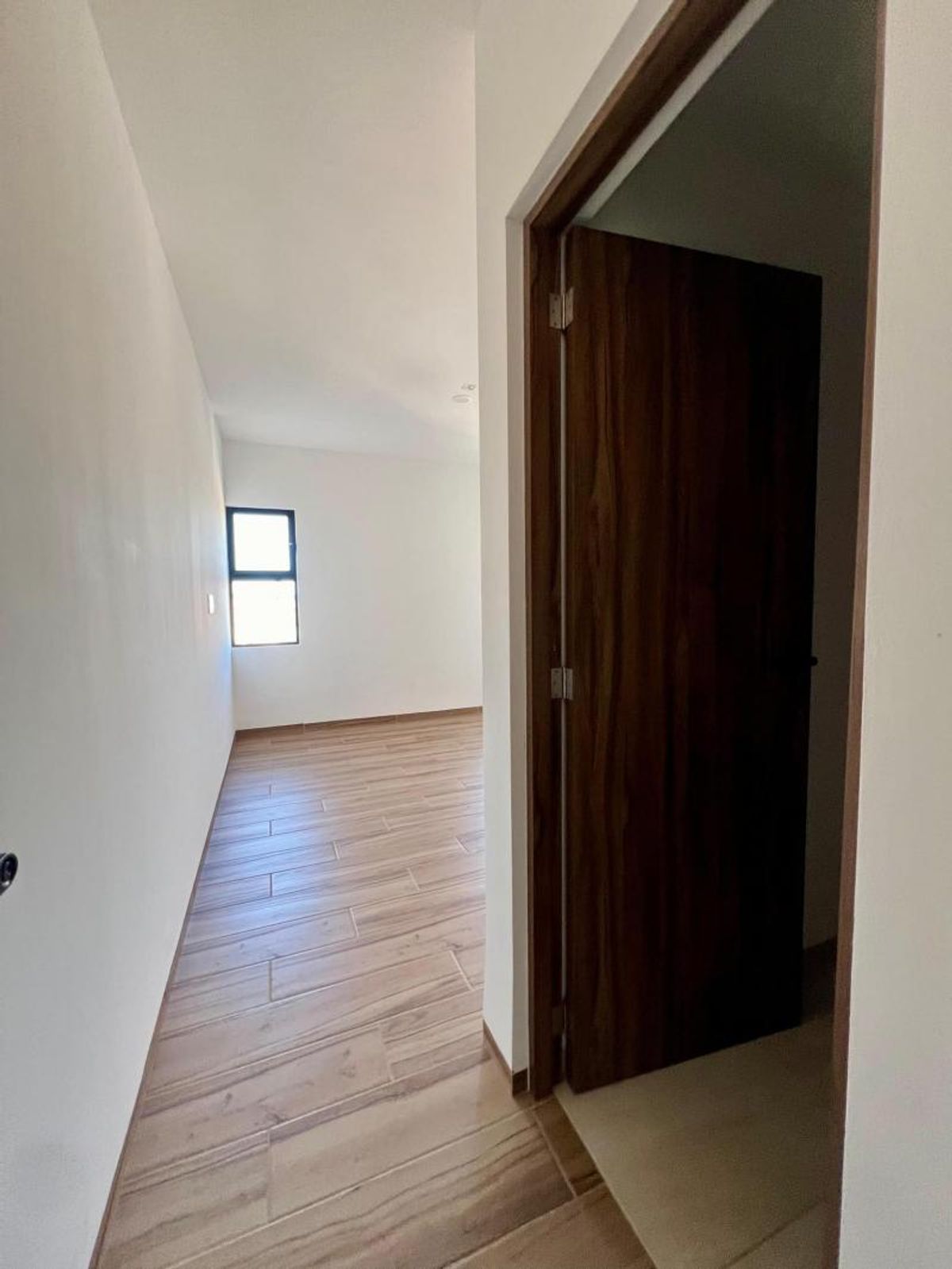 Pasillo con piso tipo madera, paredes blancas, ventana al fondo y puerta de madera