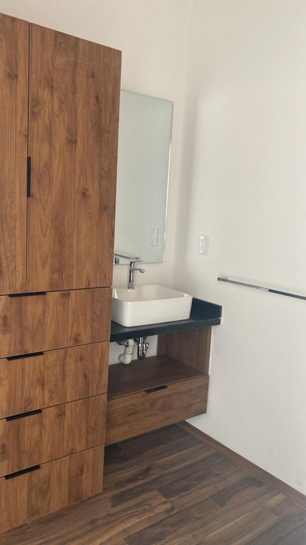 Baño con armario alto de madera, lavabo blanco sobre cubierta negra y espejo vertical