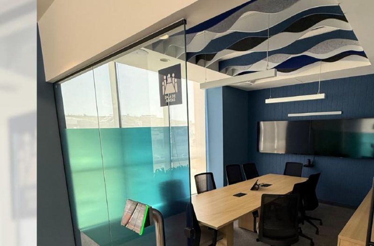 Sala de juntas con mesa de madera, sillas negras, muro de cristal y plafón azul