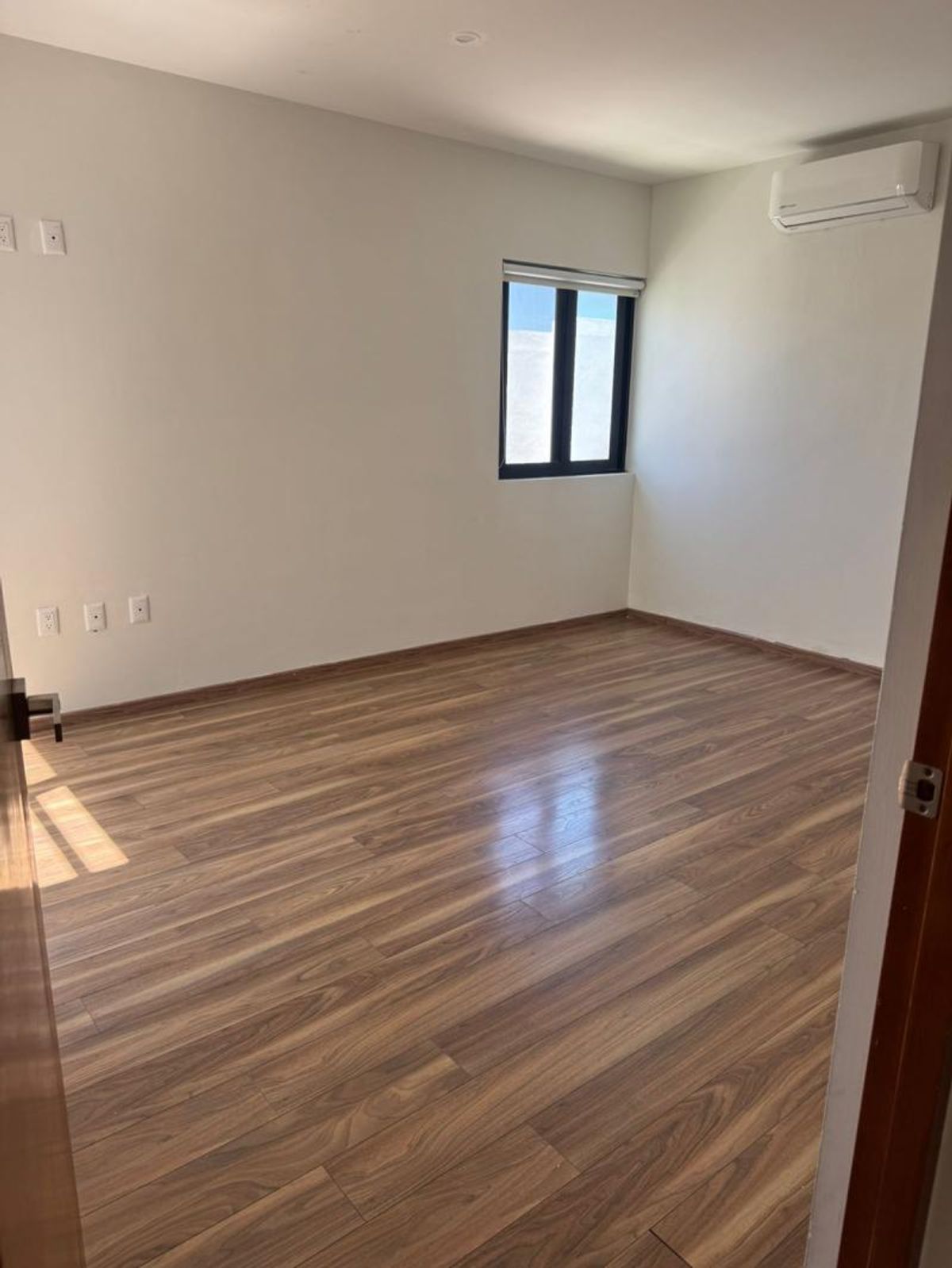 Recámara vacía con piso laminado tipo madera, paredes claras, ventana y aire acondicionado