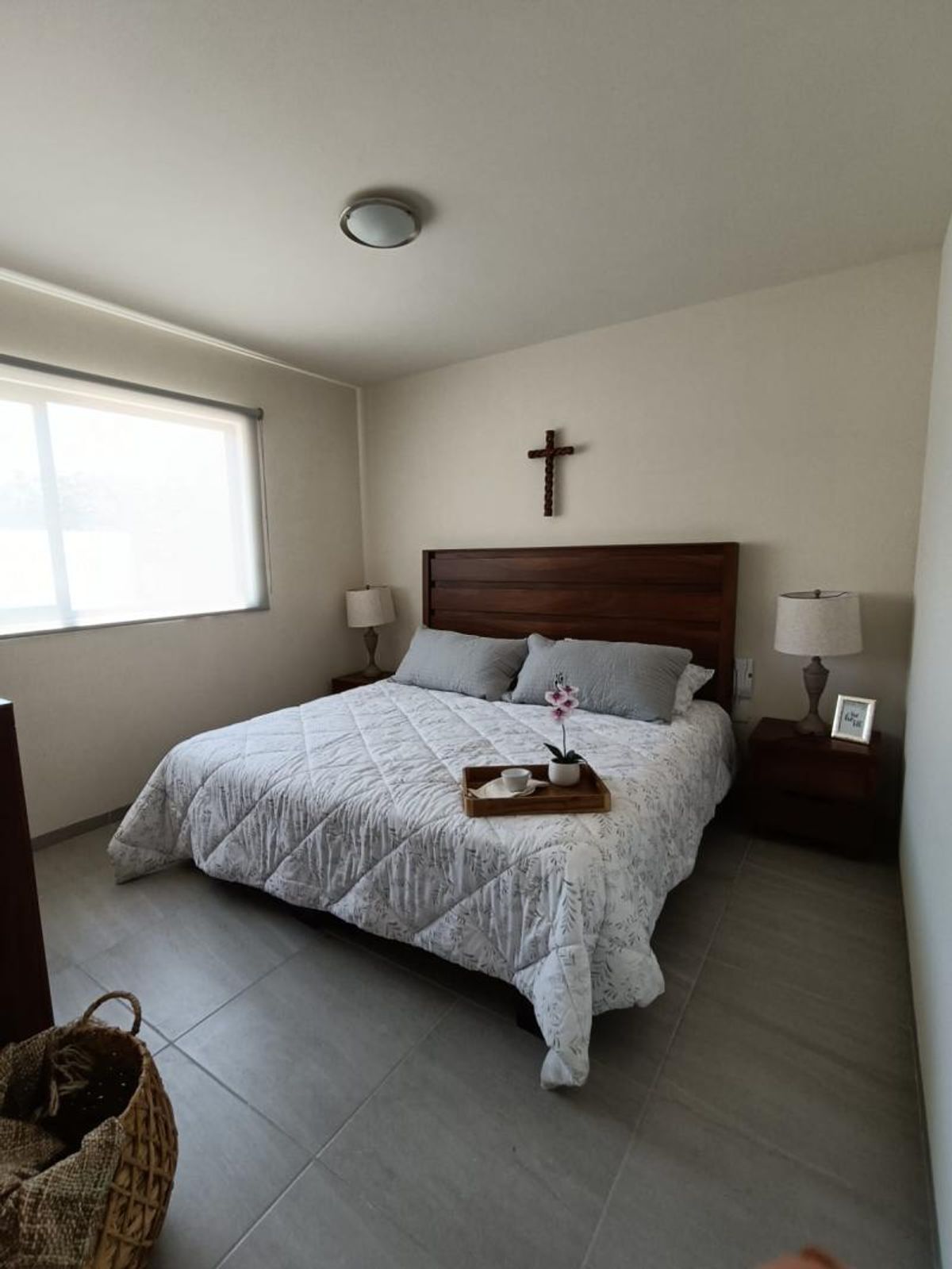 Recámara con cama, cabecera de madera, burós con lámparas y piso de loseta gris