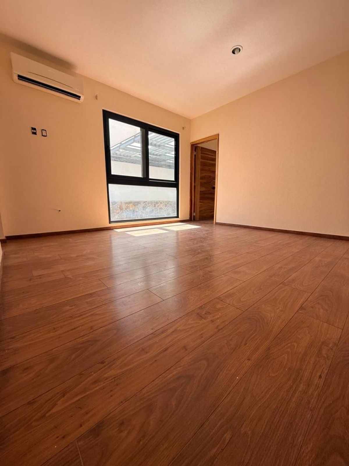 Habitación vacía con piso tipo madera, paredes beige, ventana con marco negro y aire acondicionado