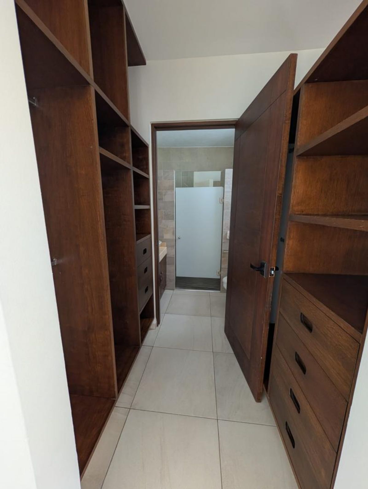 Vestidor con cajoneras y repisas de madera oscura con piso claro junto al baño