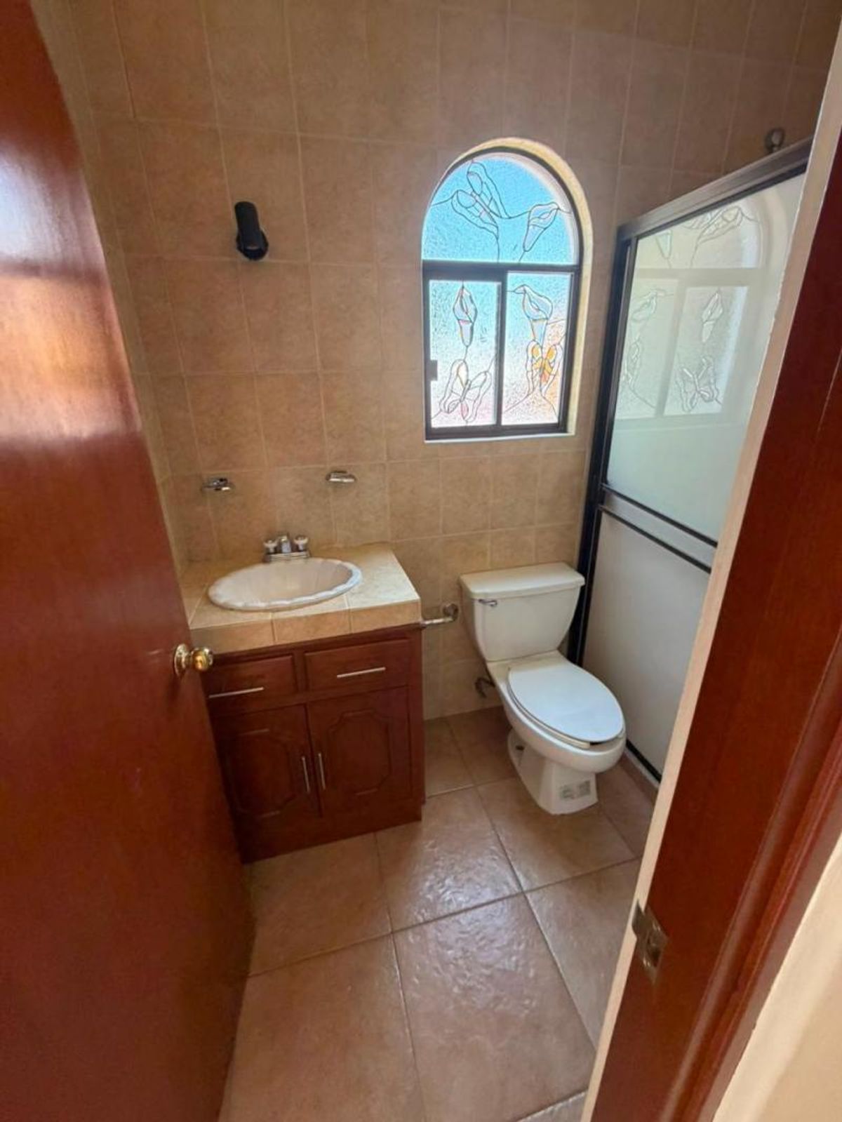 Baño con paredes de azulejo, mueble de madera, inodoro blanco y ventana con arco